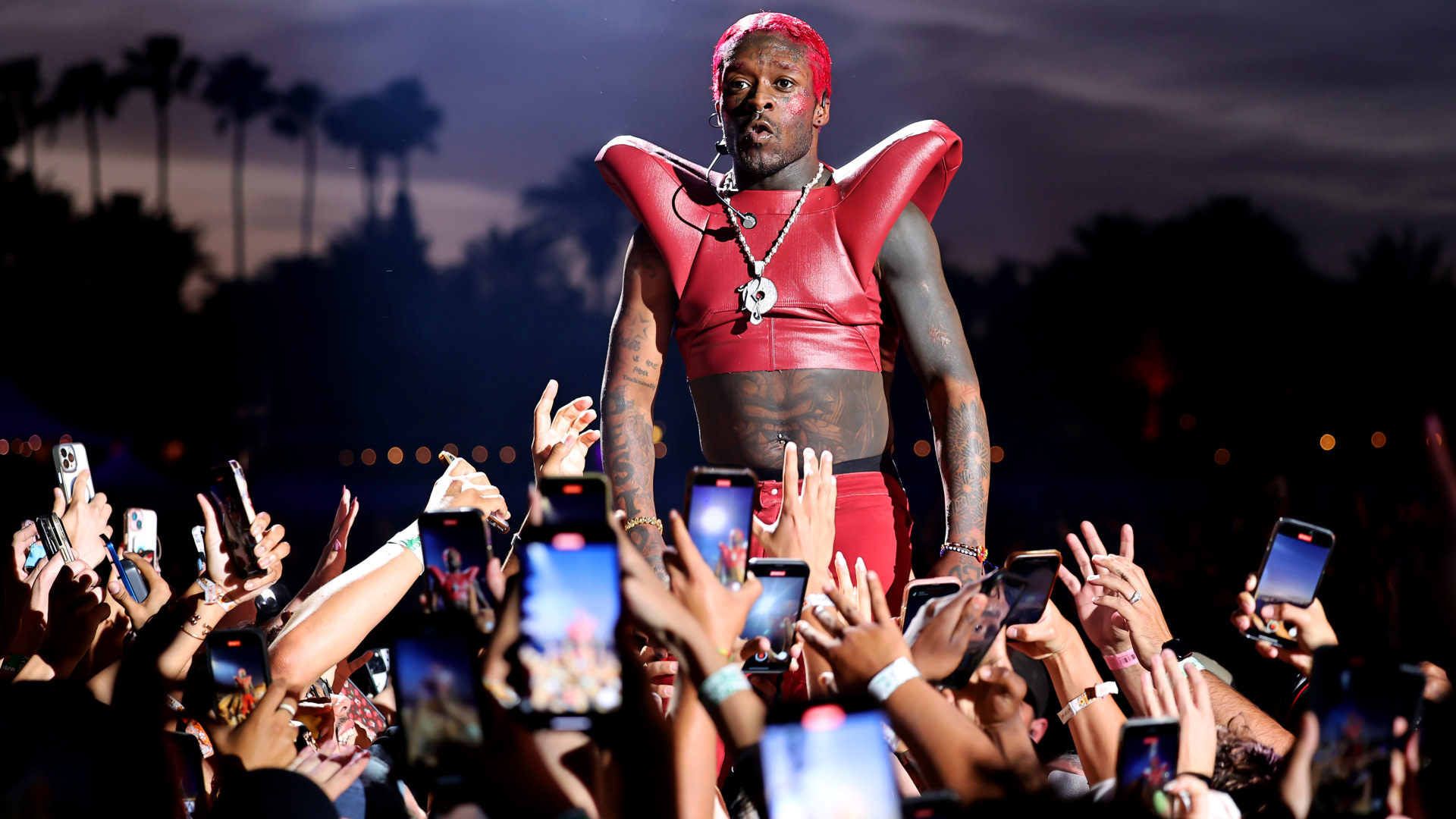 Rapper Lil Uzi Vert performing on stage, wearing a red outfit, surrounded by fans holding up phones, with a palm tree silhouette in the background.