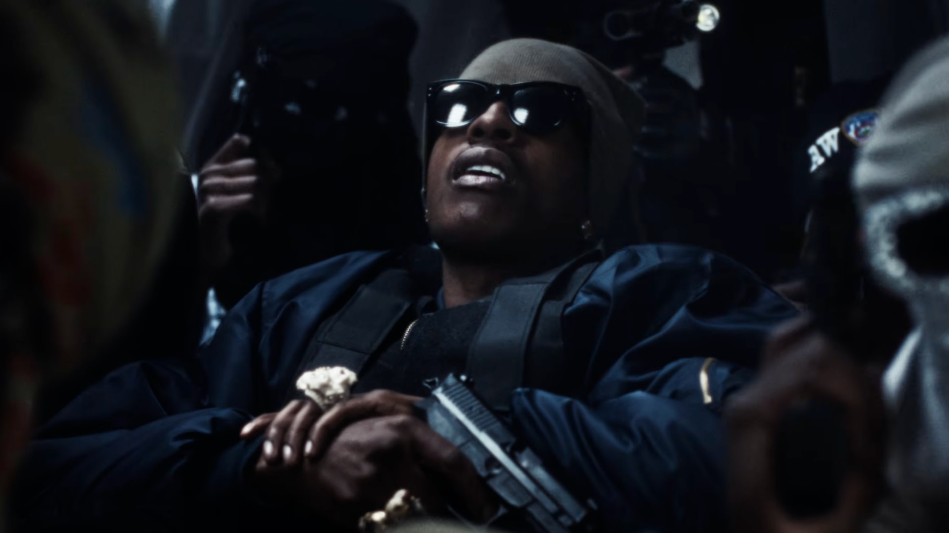 ASAP Rocky wearing sunglasses and a beanie, holding a gun, surrounded by others in a dark setting.