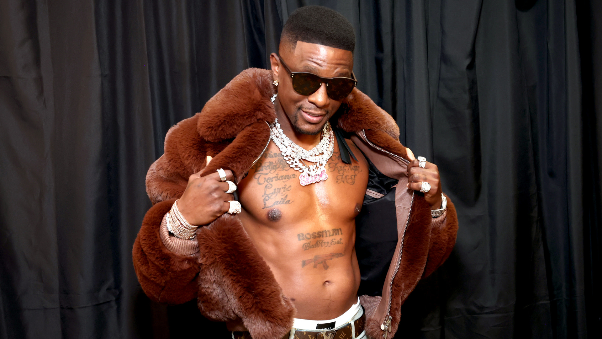Boosie Badazz in a fur coat and sunglasses, showing tattoos on his chest. Black background.