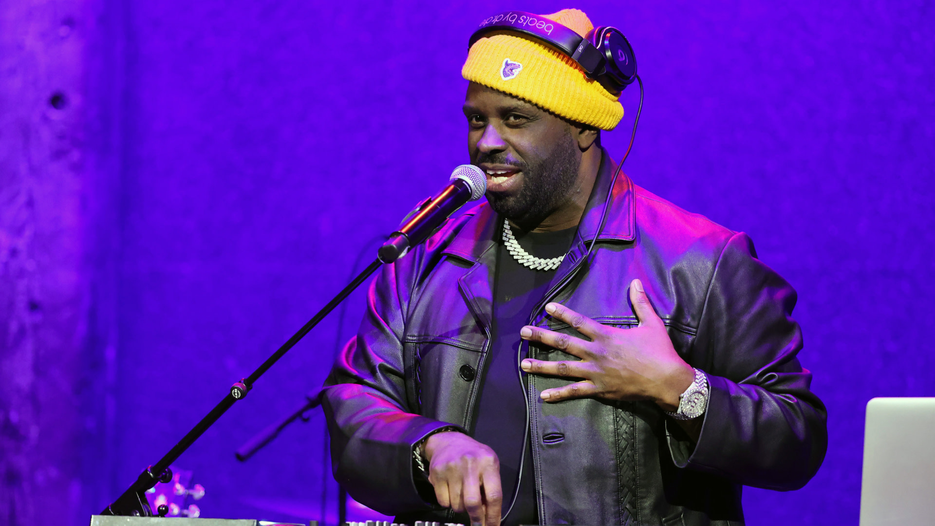 Funk Flex wearing a yellow beanie and headphones performs on stage with a microphone, dressed in a leather jacket.