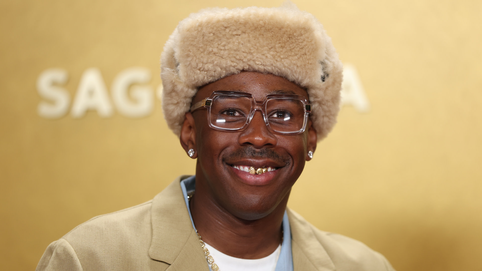 Tyler, The Creator wearing a fur hat, large glasses, and a beige suit, smiling with a gold grill against a gold background.