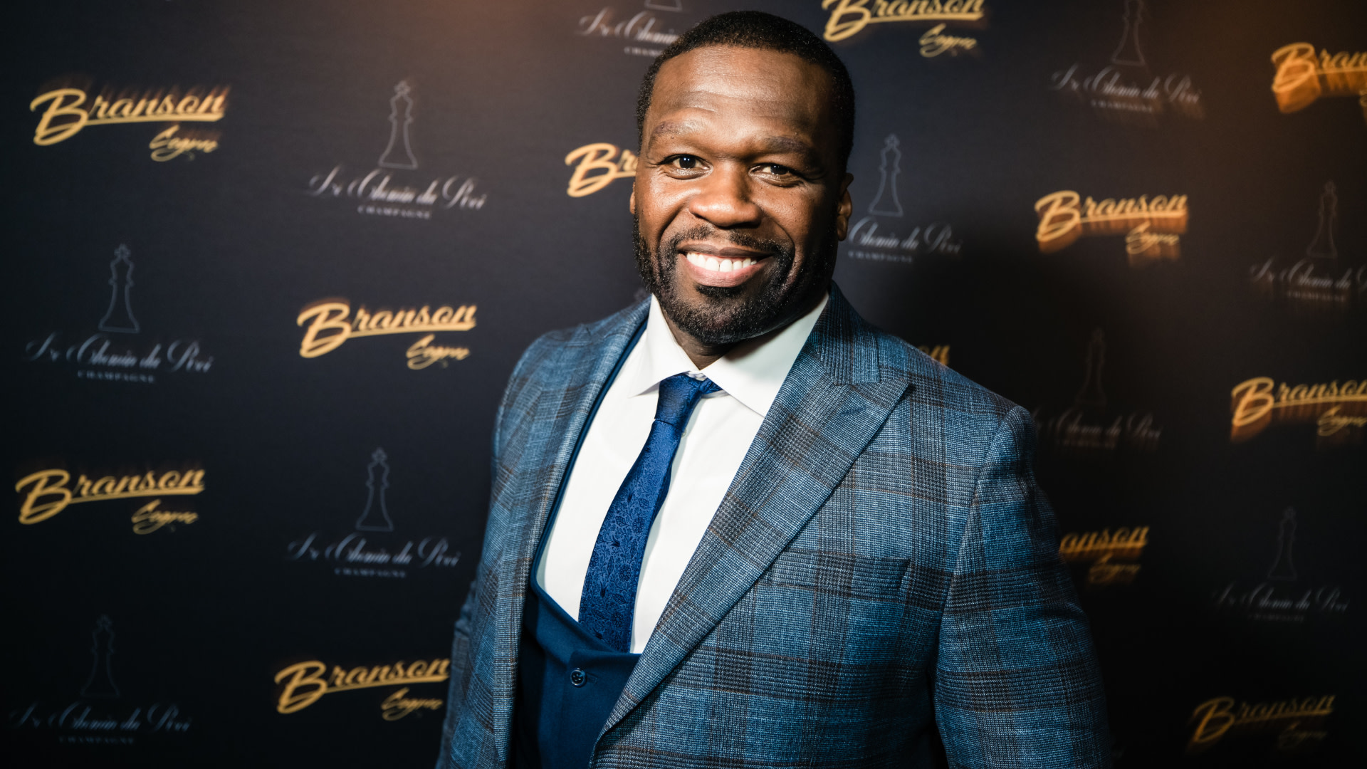 50 Cent, wearing a blue plaid suit and tie, smiling at an event with a Branson Cognac backdrop.