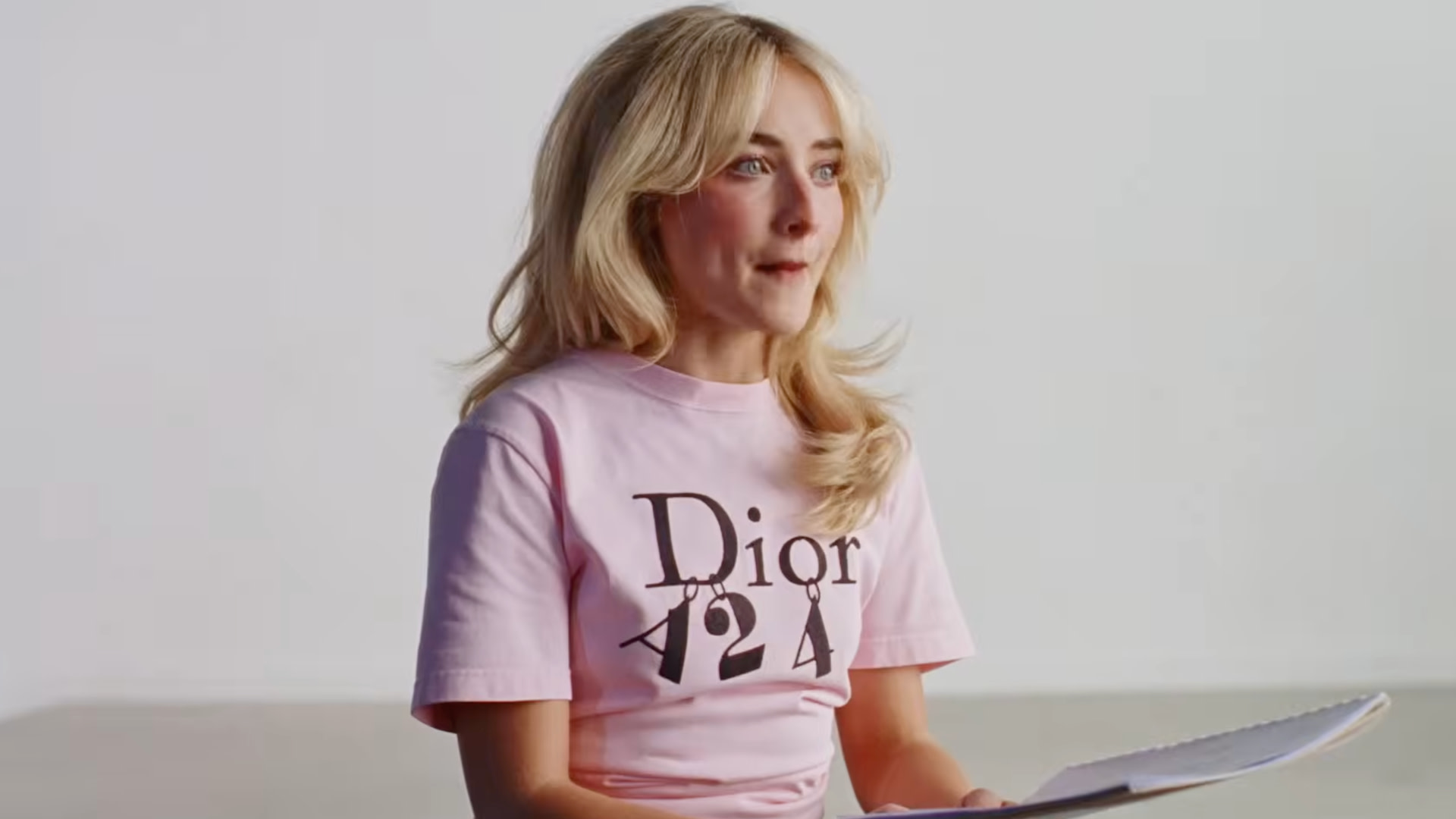 Sabrina Carpenter with long blonde hair, wearing a pink Dior A24 shirt, is sitting and holding a notebook.