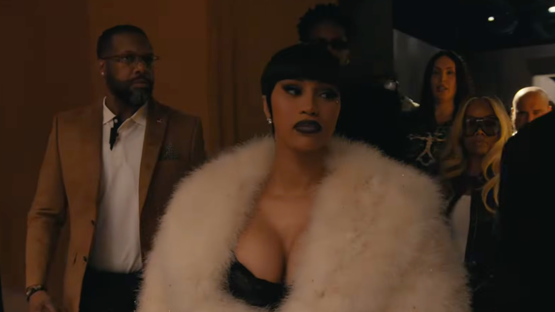 Cardi B in a glamorous outfit with a white fur coat, surrounded by people in a dimly lit setting.