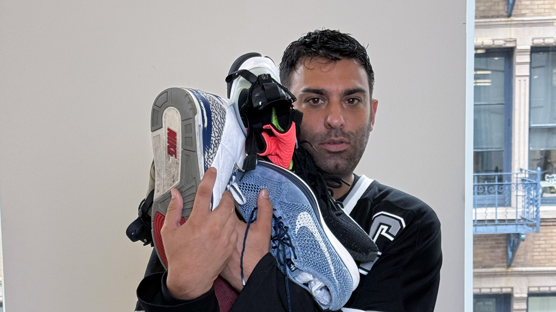 A man holding several pairs of sneakers, including Nike shoes, in front of a window with a view of a building.