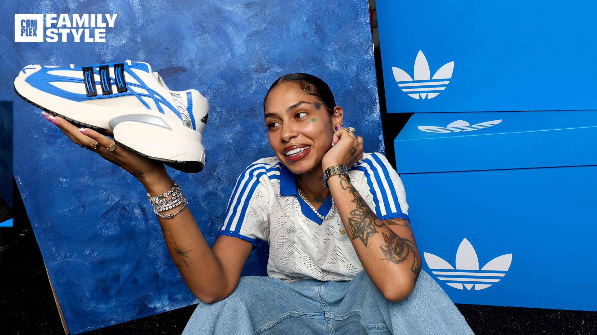 A person holding a blue and white Adidas sneaker, sitting in front of blue Adidas boxes, smiling. Wearing a striped shirt and jeans.
