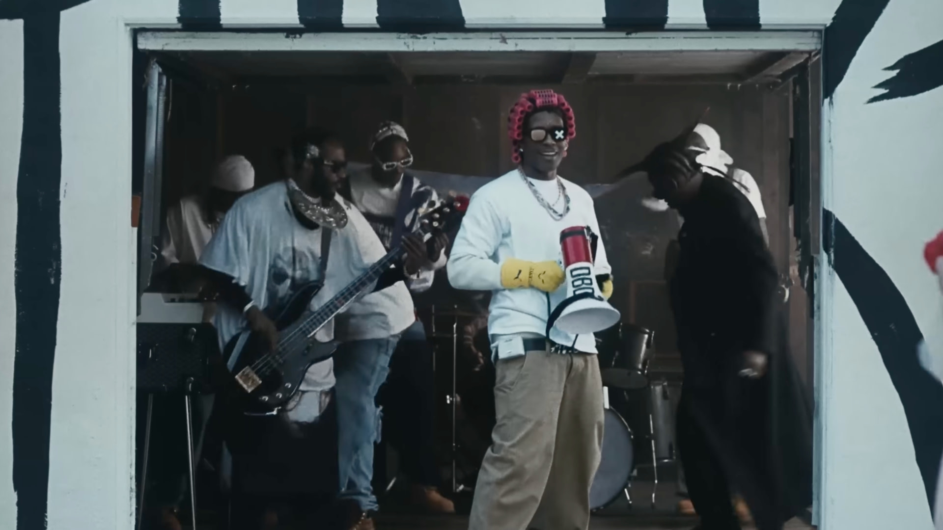 A group of musicians performing in a room. ASAP Rocky holds a megaphone, wearing sunglasses and a red hat. Instruments are visible.