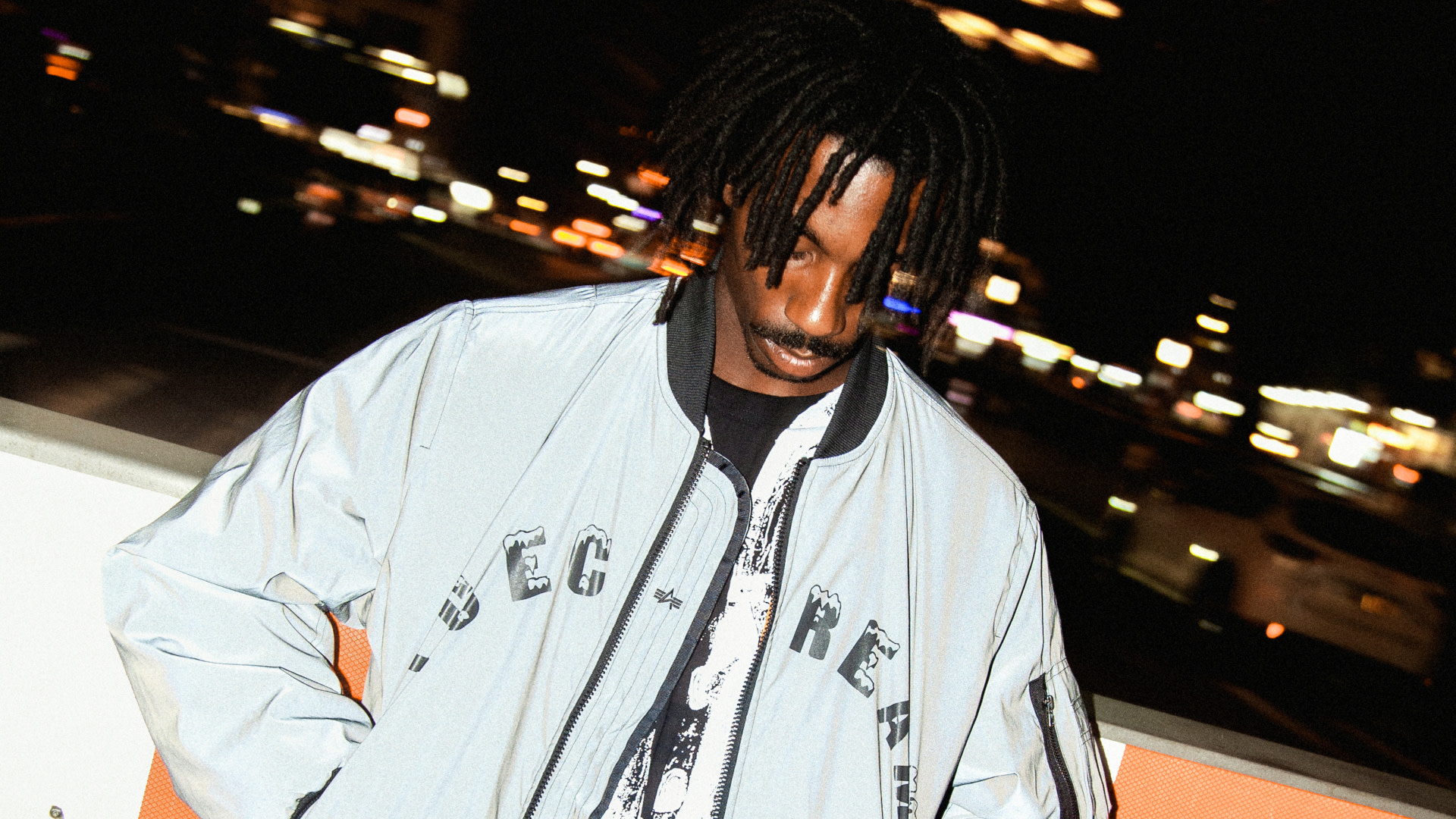 A person with dreadlocks wearing a light jacket stands in a city at night, with blurred lights in the background.