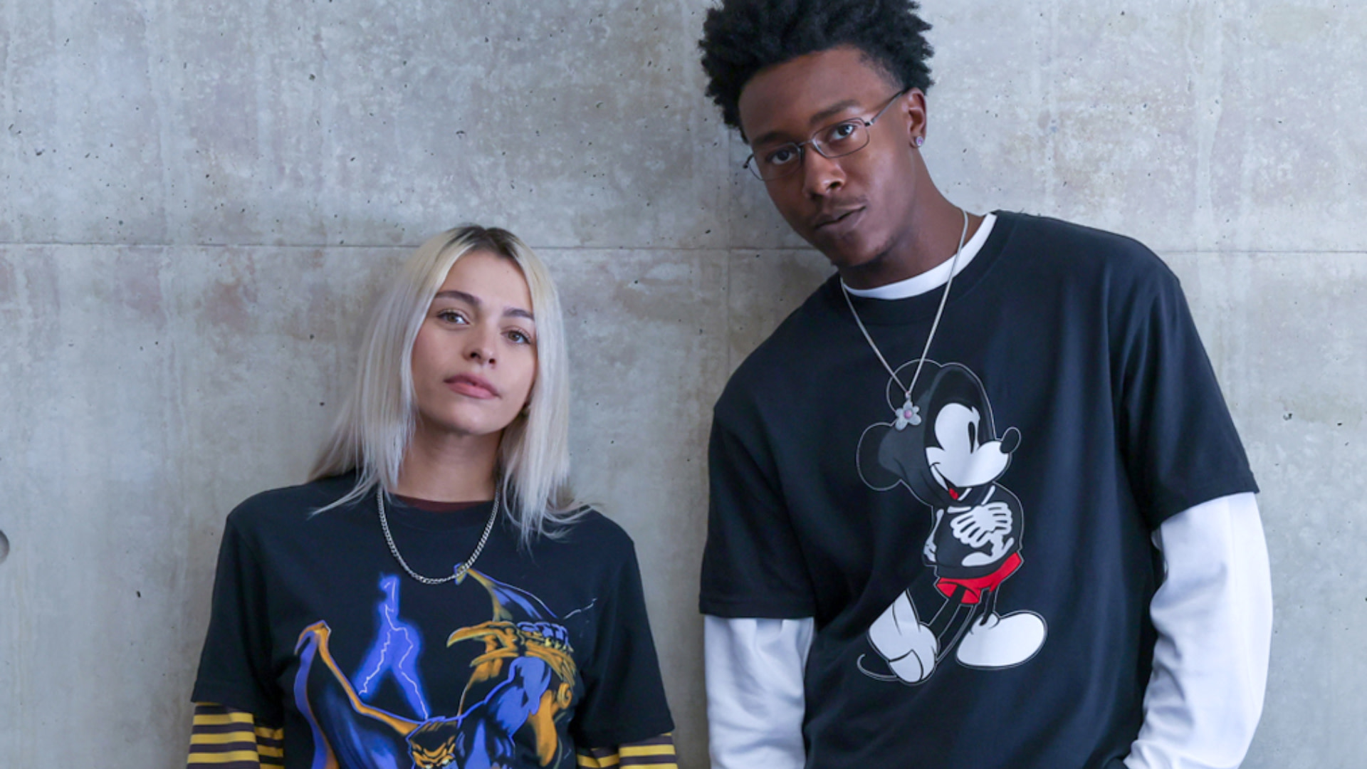 Two people posing against a concrete wall. One wears a graphic tee with a character, the other with Mickey Mouse. Casual style.