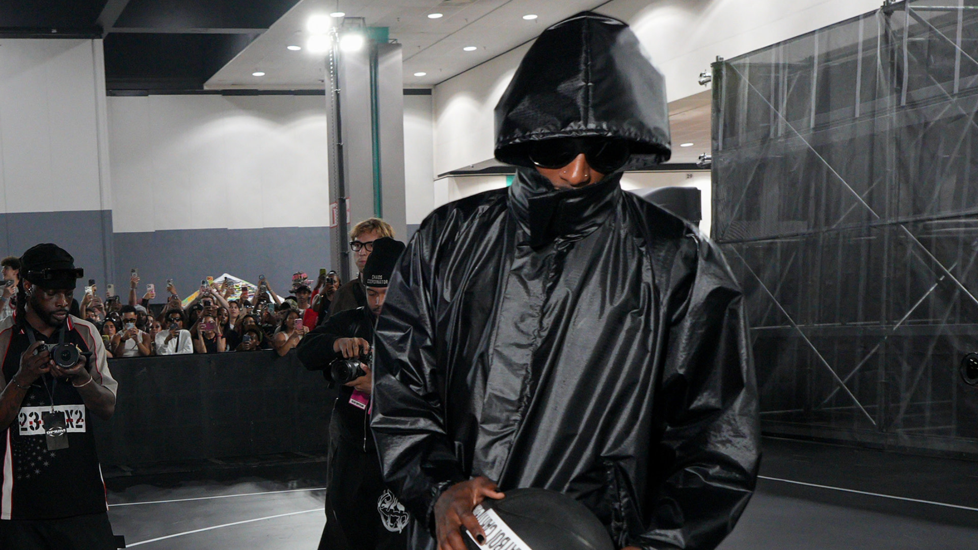 Playboi Carti in a black hooded jacket and sunglasses holds a cap, surrounded by photographers and a crowd in an indoor setting.