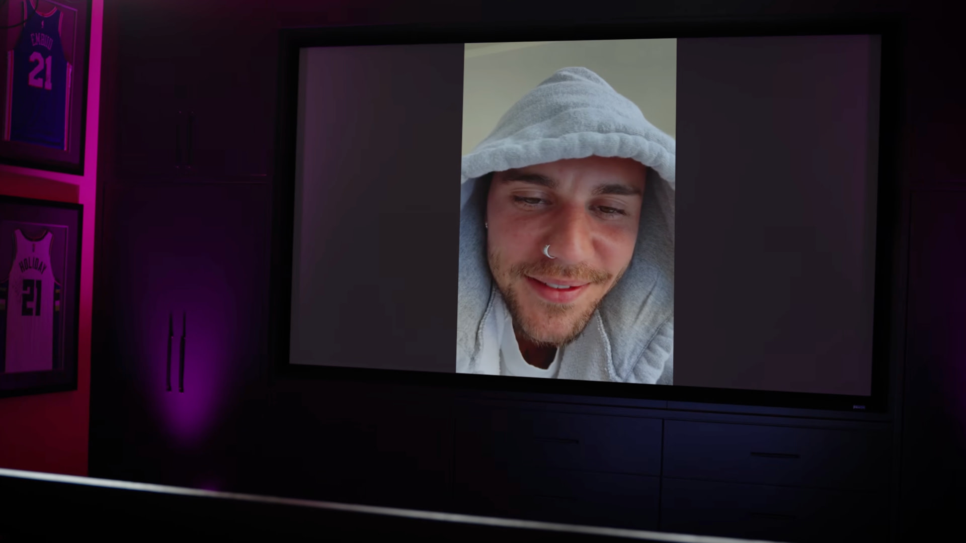 Justin Bieber in a hoodie is seen on a large screen in a dimly lit room with framed sports jerseys on the wall.