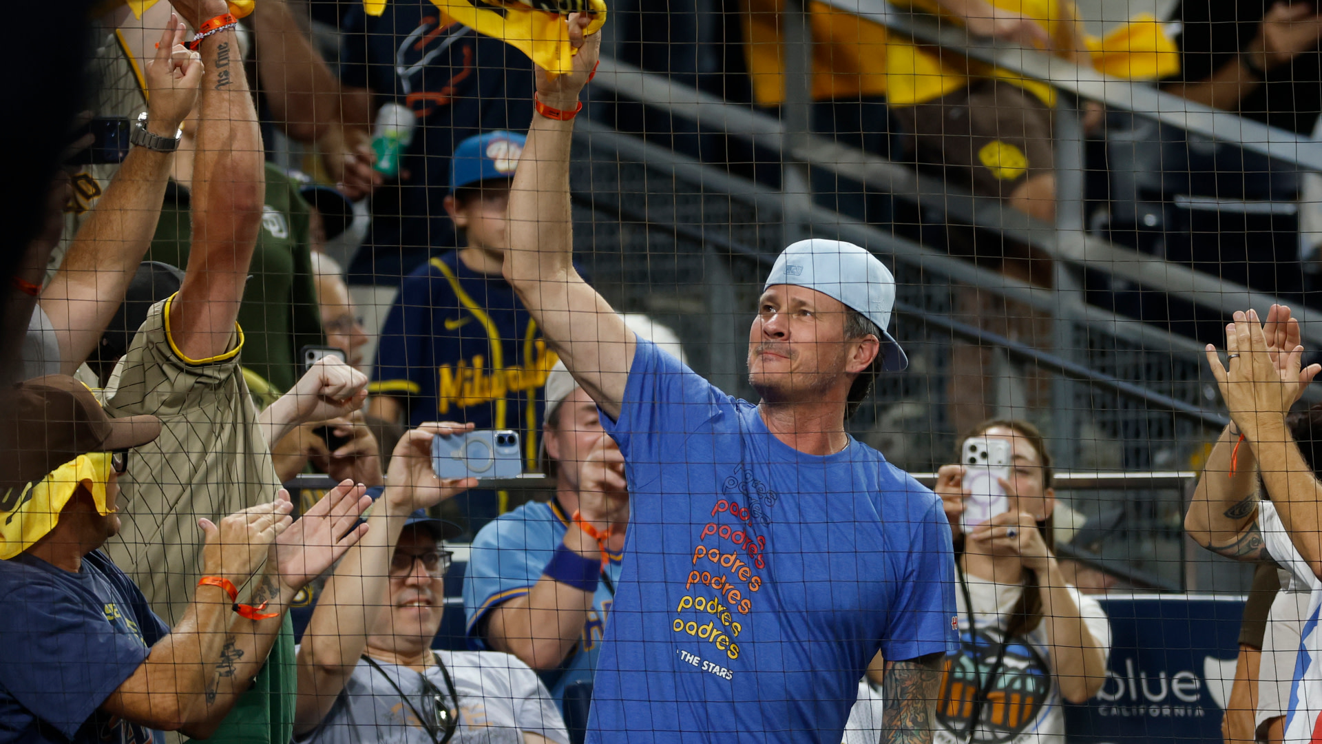 Tom DeLonge in a blue shirt and backwards cap holds up a glove at a baseball game, surrounded by applauding fans.