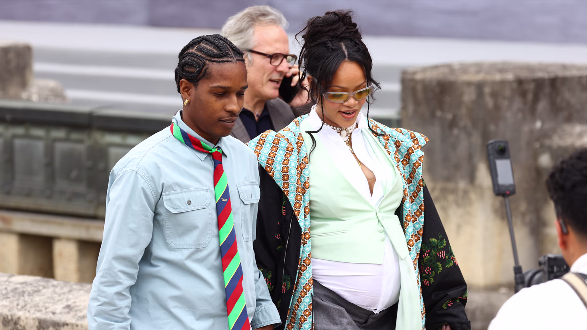 A$AP Rocky and Rihanna walking together. A$AP Rocky wears a blue shirt and striped tie, Rihanna wears a patterned coat and sunglasses.