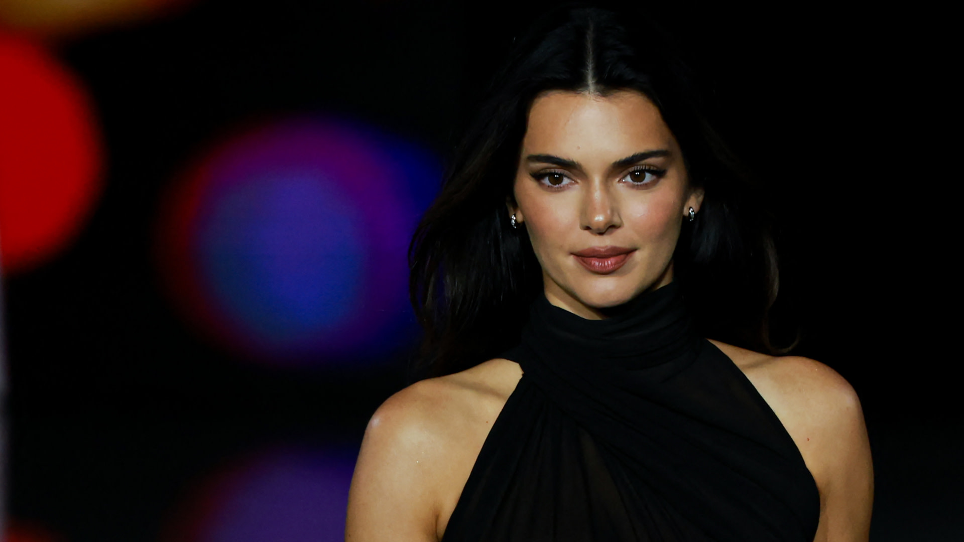 Kendall Jenner in a black halter dress, walking a runway with blurred colorful lights in the background.