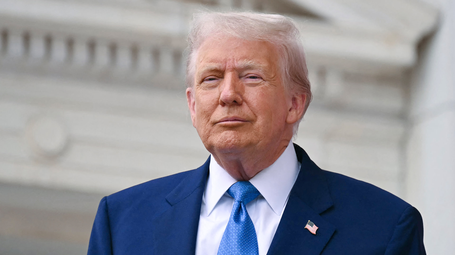 Donald Trump in a blue suit and tie stands in front of a building with white columns.
