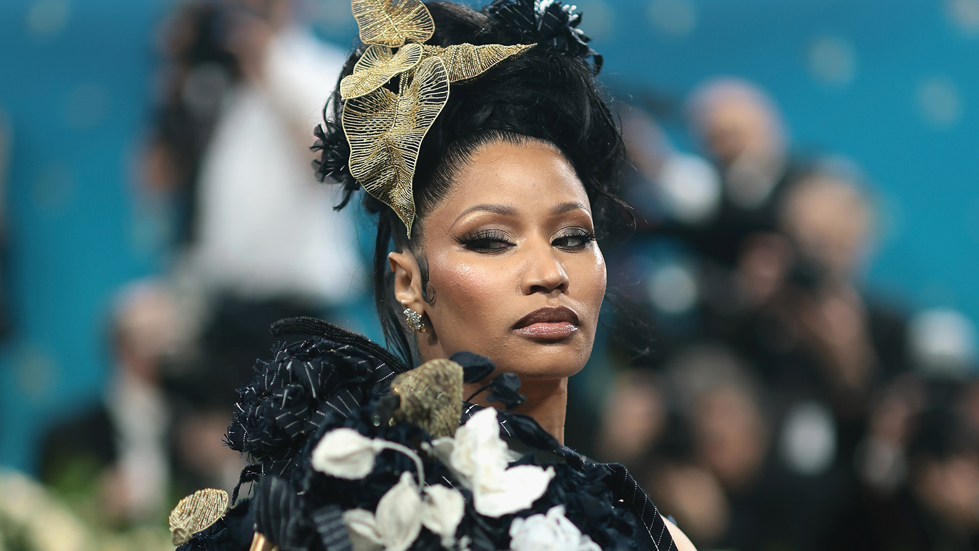 Nicki Minaj at a glamorous event, wearing an elaborate black and gold outfit with floral accents and a striking headpiece.