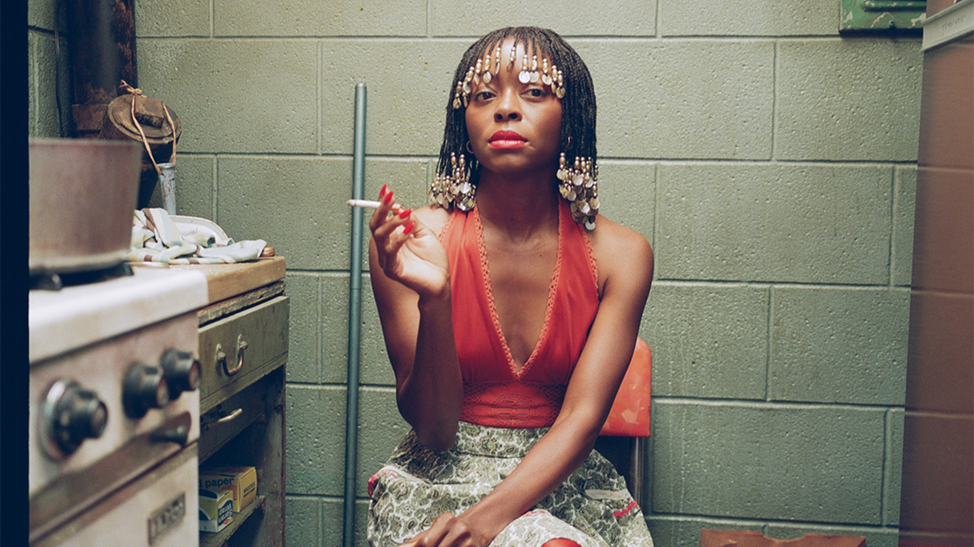 A woman in a red top and beaded hair sits in a kitchen, holding a cigarette, with a confident expression.
