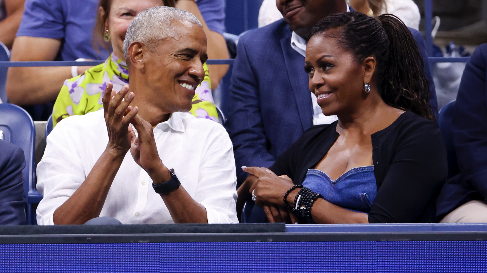 Barack Obama and Michelle Obama smiling and interacting at a public event, seated in a crowd.