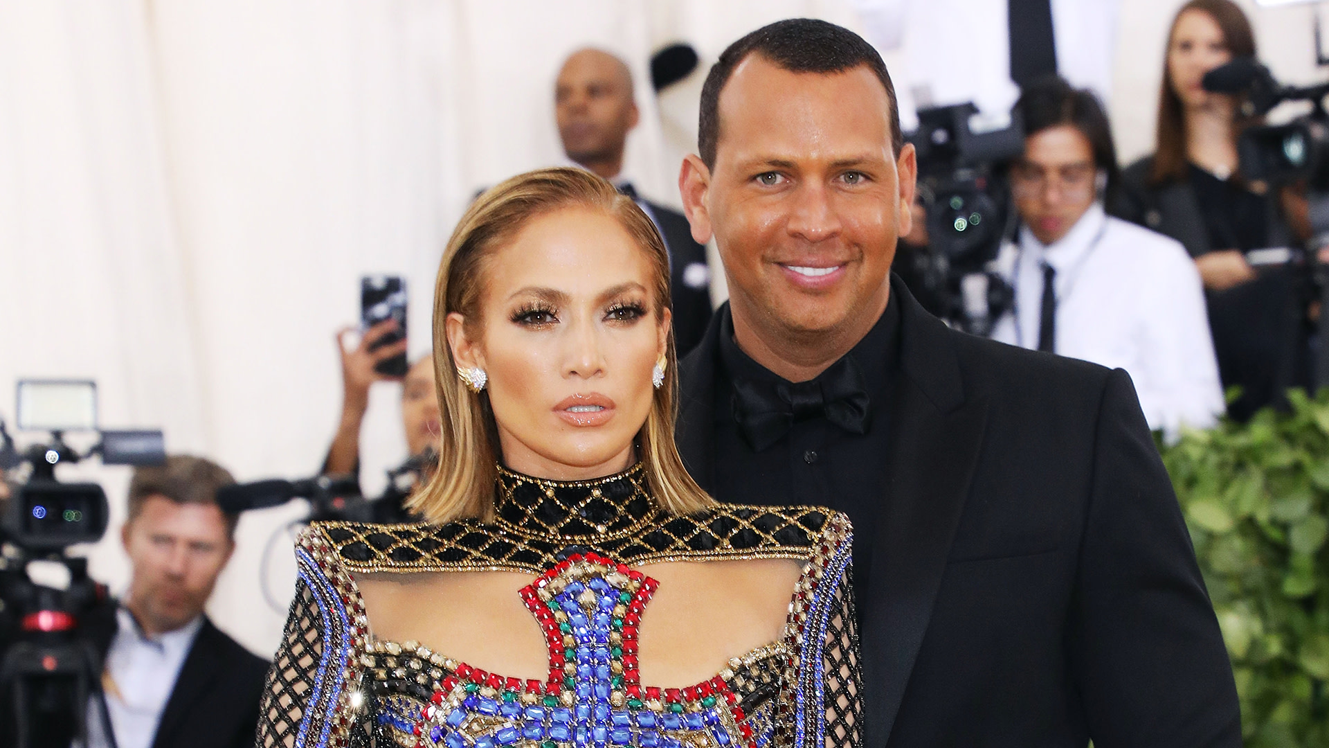 Jennifer Lopez in a jeweled gown and Alex Rodriguez in a suit pose at an event with photographers in the background.