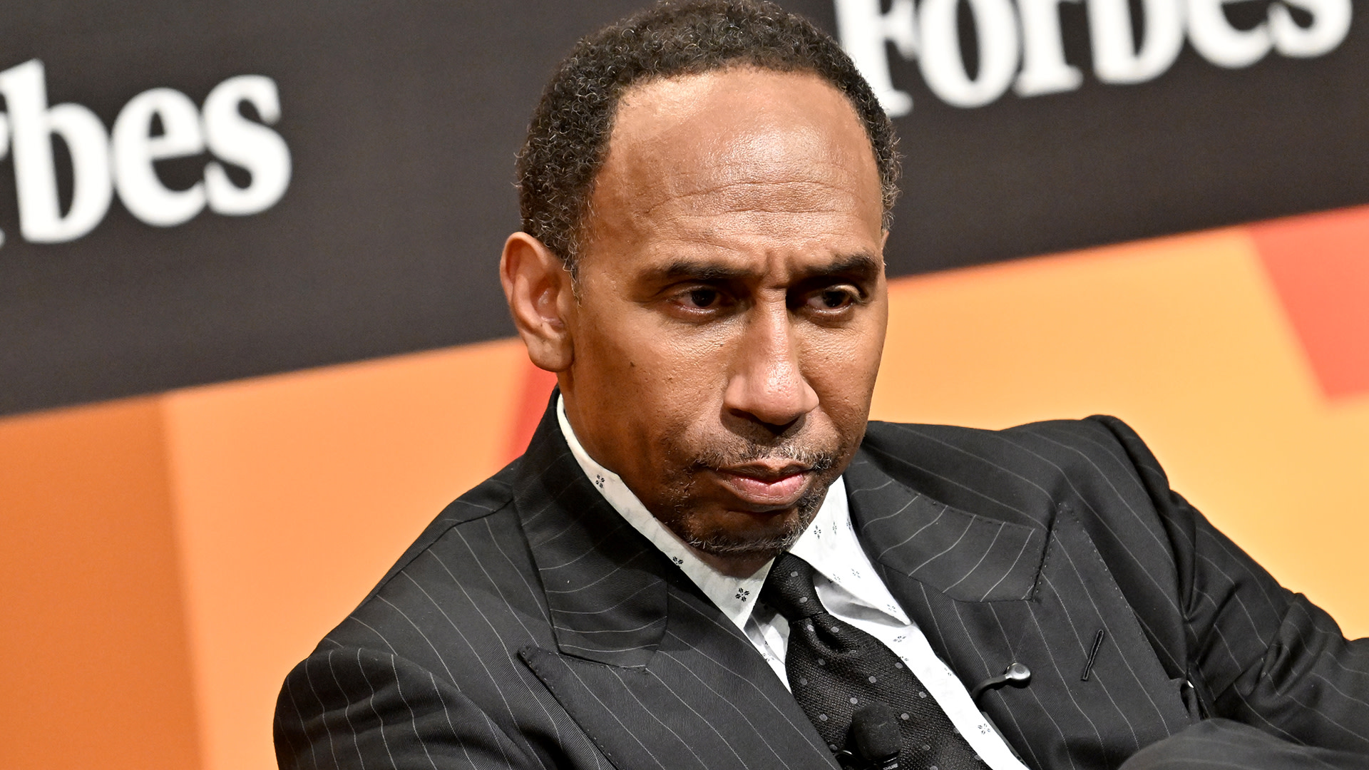 Stephen A. Smith in a pinstripe suit sits pensively against a backdrop with the Forbes logo.