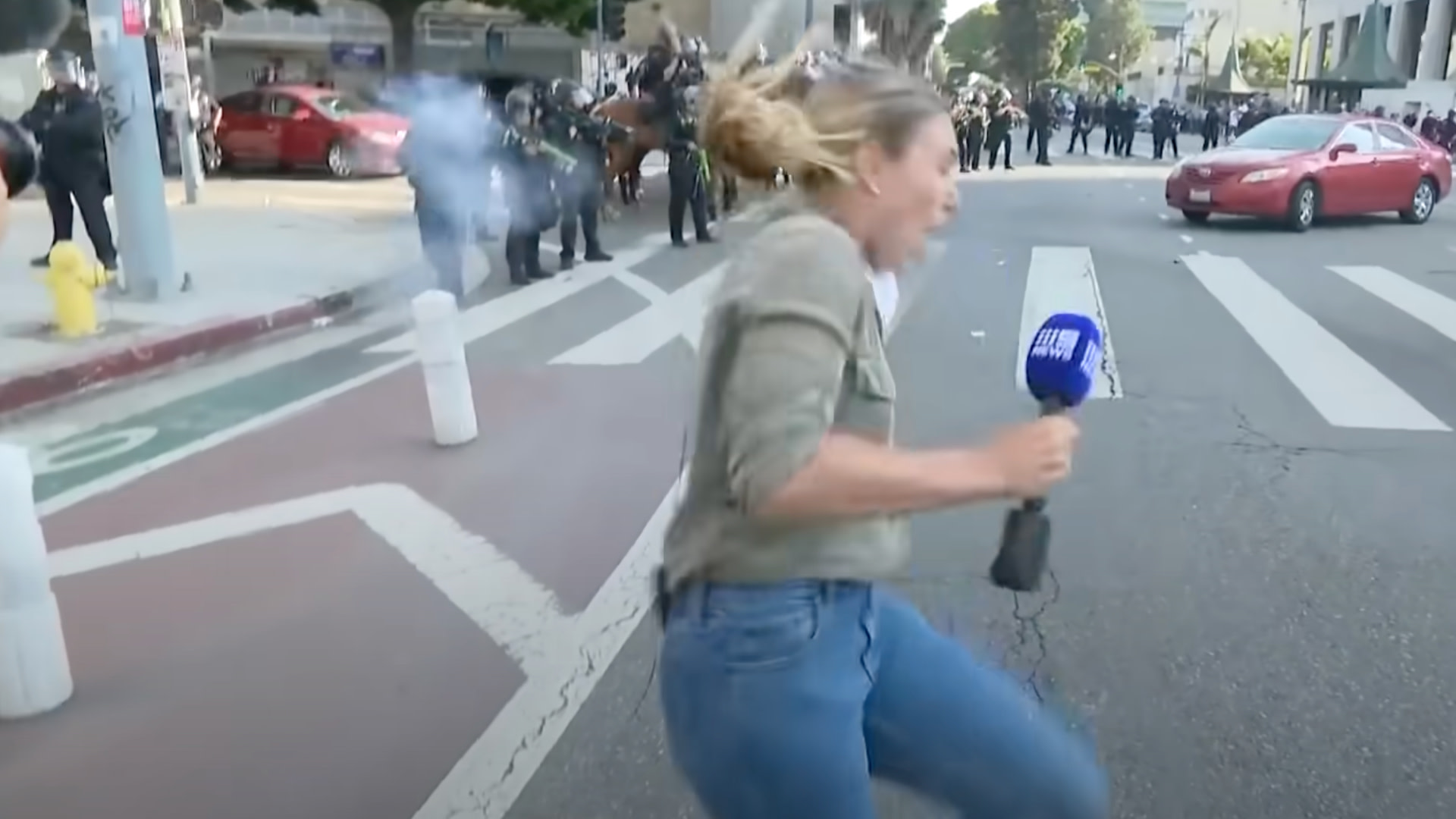 A reporter reacts while holding a microphone during a protest. Police officers and smoke are visible in the background on a city street.