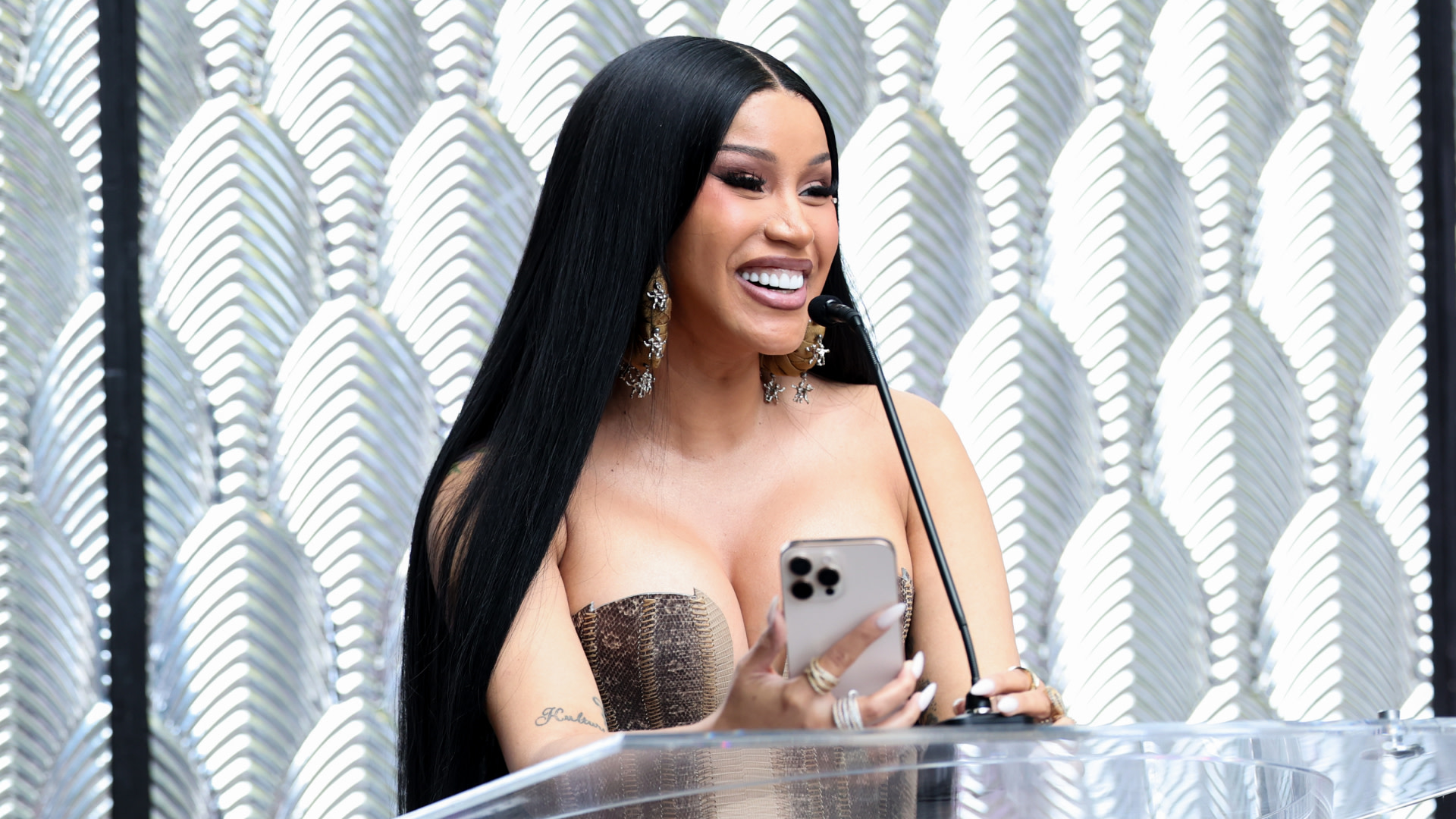 Cardi B, in a strapless dress, smiles while holding a phone at a podium, with an elegant patterned background.