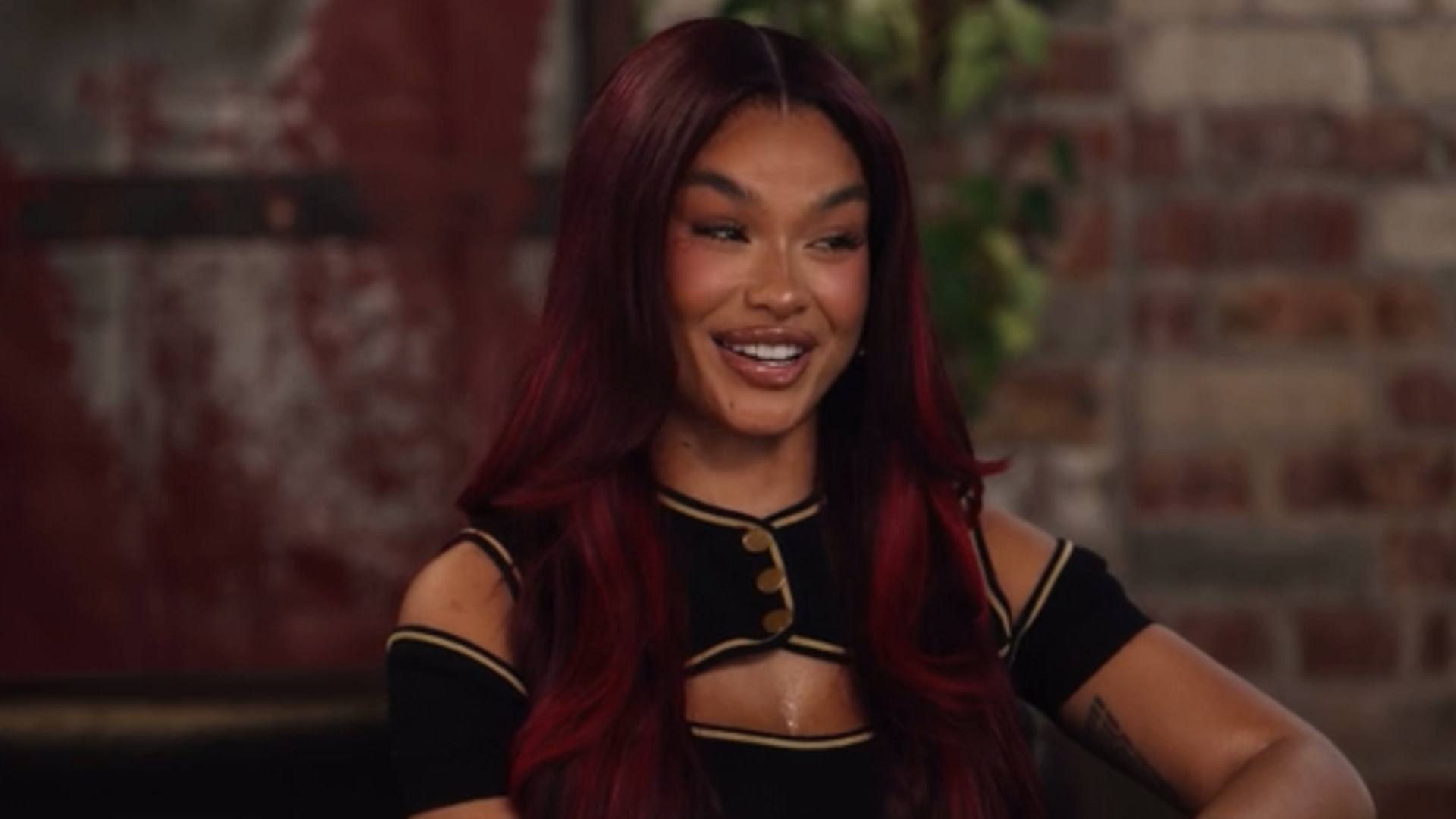 India Love with long red hair smiles, sitting against a brick wall background. She wears a black top with gold accents.