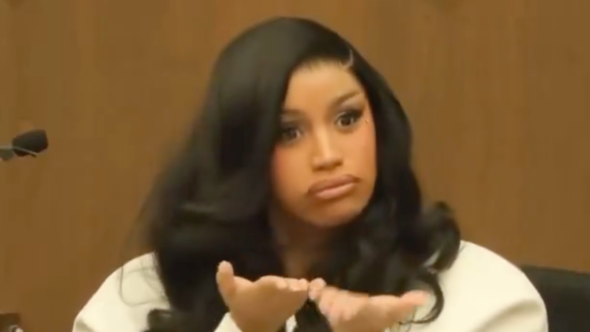 Cardi B in a courtroom, making a confused gesture with her hands, wearing a white outfit and long wavy hair.