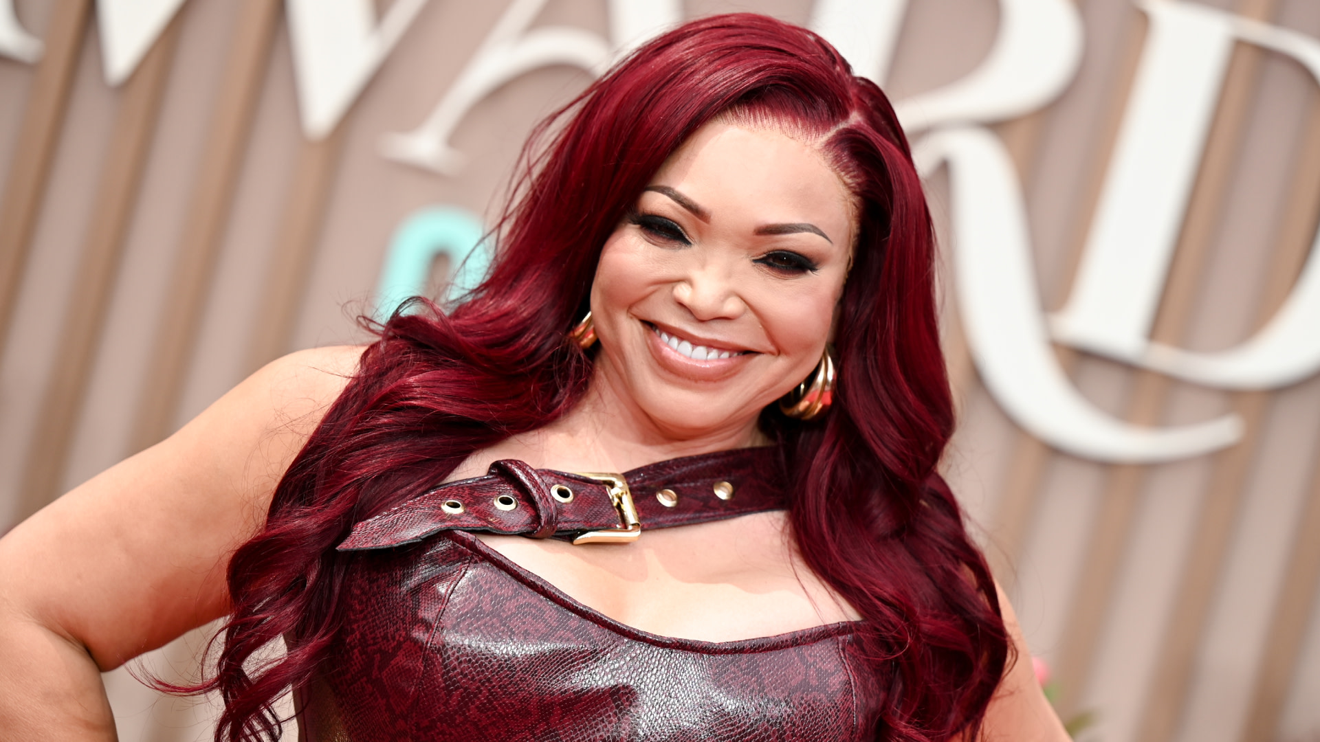 Tisha Campbell with long red hair and a leather outfit smiles at an event, with a blurred background.