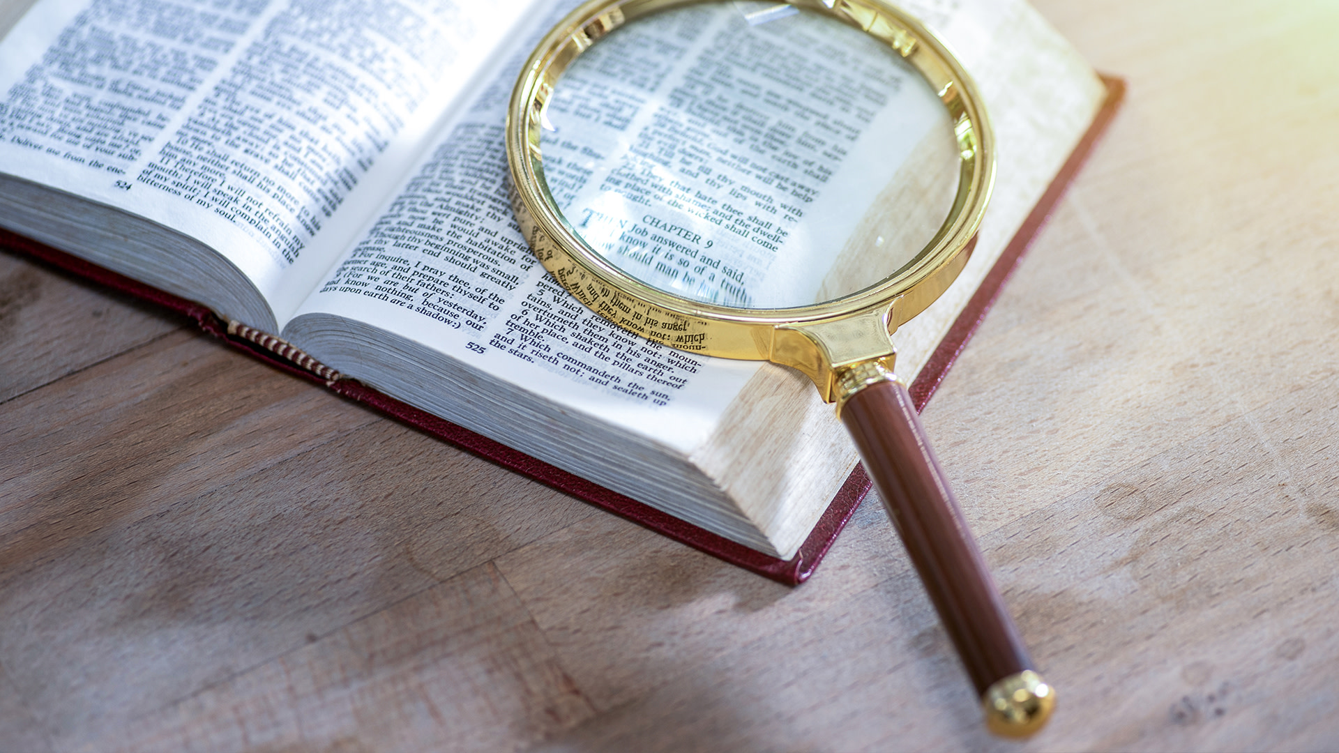 An open book with a magnifying glass resting on it, highlighting the text. The book is on a wooden surface.
