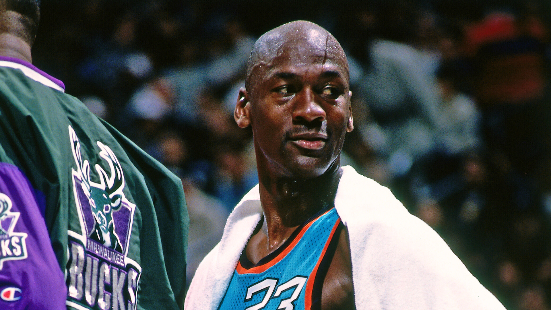 Michael Jordan, in a teal basketball jersey with the number 23, draped in a white towel, stands near a Milwaukee Bucks player.
