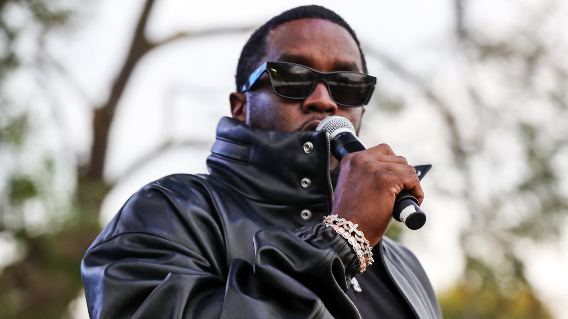 Diddy in a black leather jacket and sunglasses holding a microphone, set outdoors with blurred trees in the background.