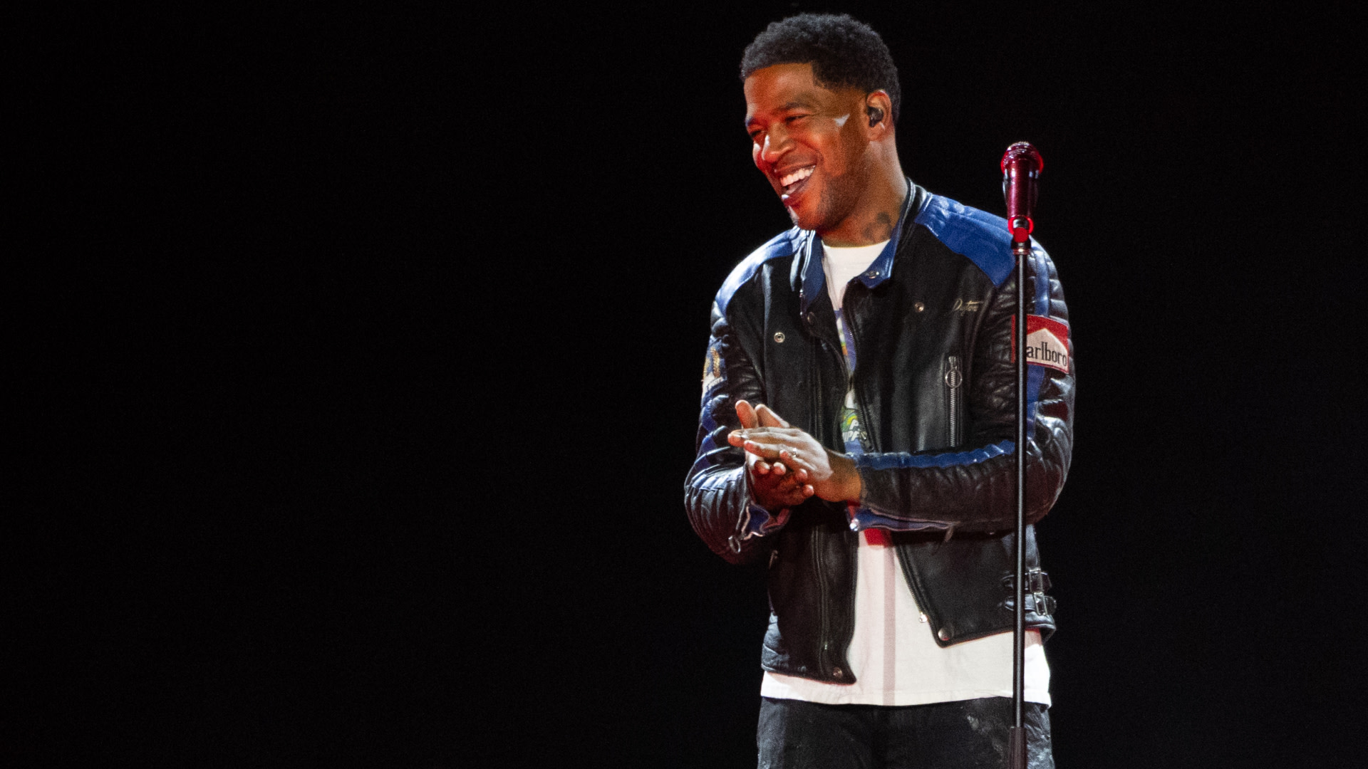 Kid Cudi on stage, wearing a black leather jacket, smiling and clapping with a microphone stand nearby.