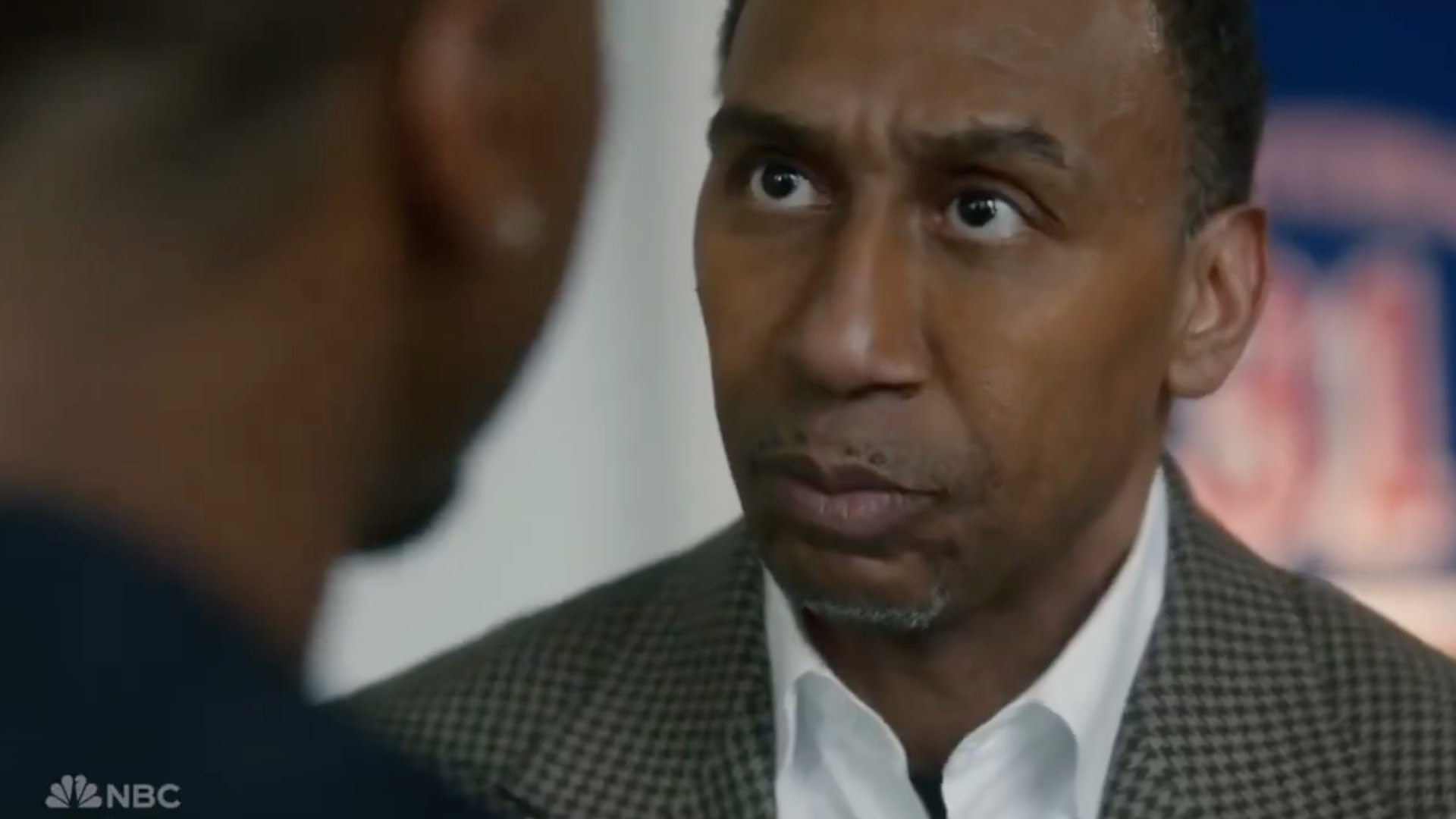 A man with a serious expression, wearing a checkered blazer and white shirt, faces another person in a close-up shot.