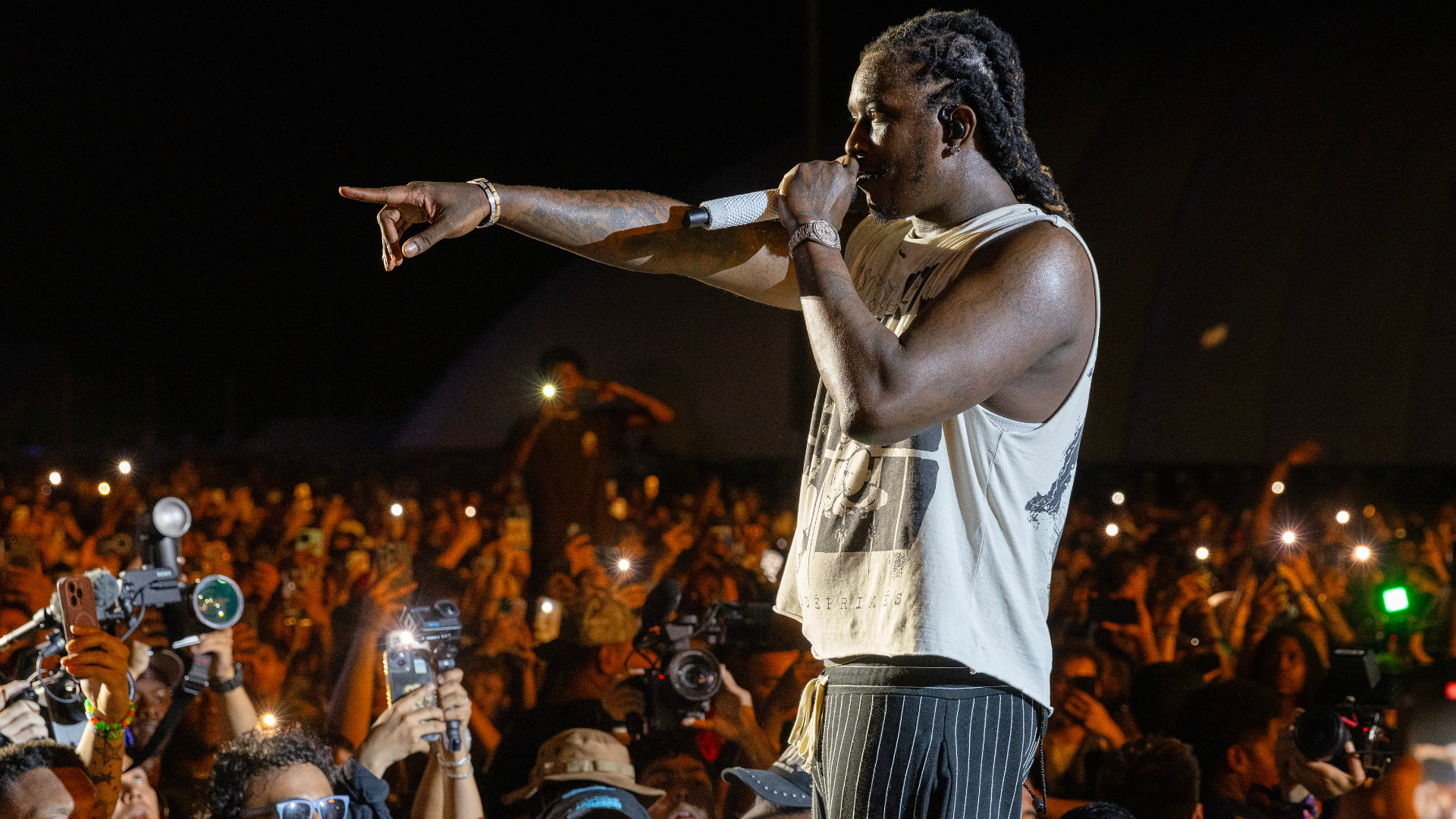 Young Thug with braided hair sings into a microphone on stage, pointing towards the crowd. The audience holds up phones and cameras.