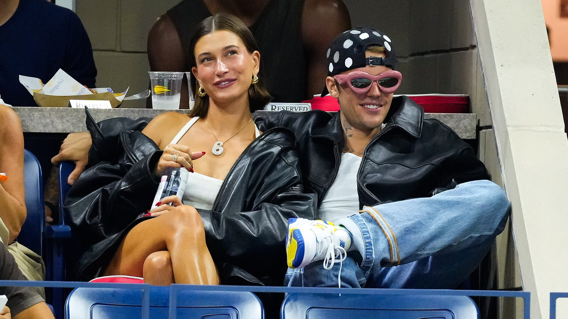Hailey Bieber and Justin Bieber are sitting together at an event, wearing black jackets. Hailey has a drink, and Justin wears sunglasses.