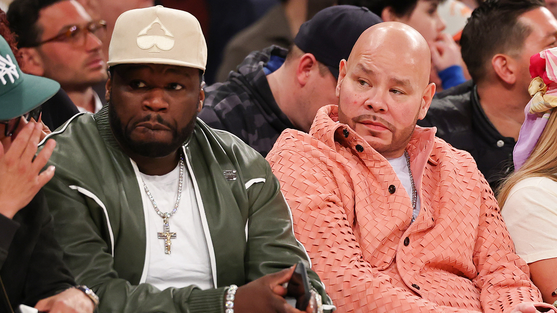 50 Cent in a green jacket and cap sits next to Fat Joe in an orange jacket at a sports event, surrounded by other spectators.
