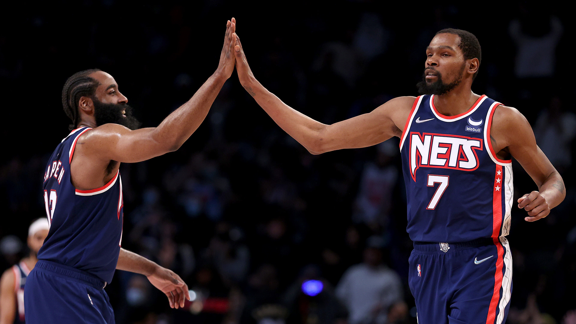James Harden and Kevin Durant of the Brooklyn Nets high-five on the basketball court, wearing navy jerseys with the Nets logo.