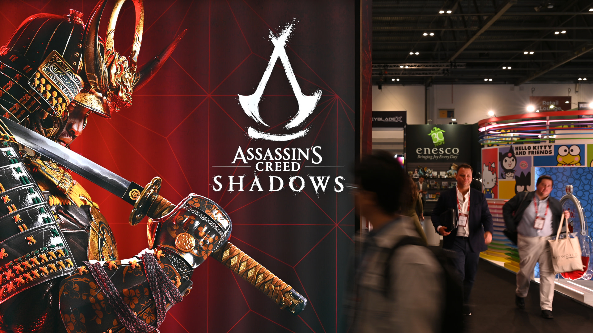 A large "Assassin's Creed: Shadows" poster featuring a samurai warrior. People walk by at a convention with colorful booths.