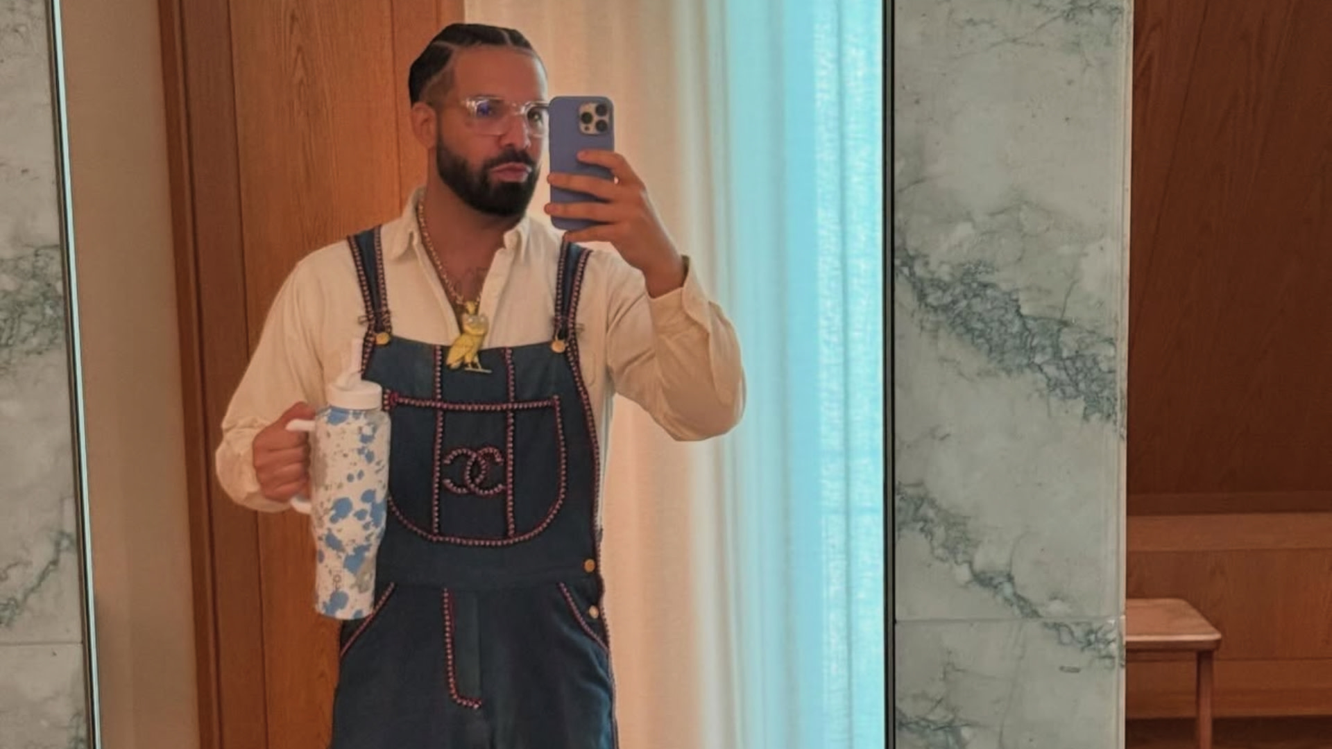 Drake with braided hair takes a mirror selfie wearing glasses, a beige shirt, and denim overalls with a Chanel logo, holding a patterned cup.