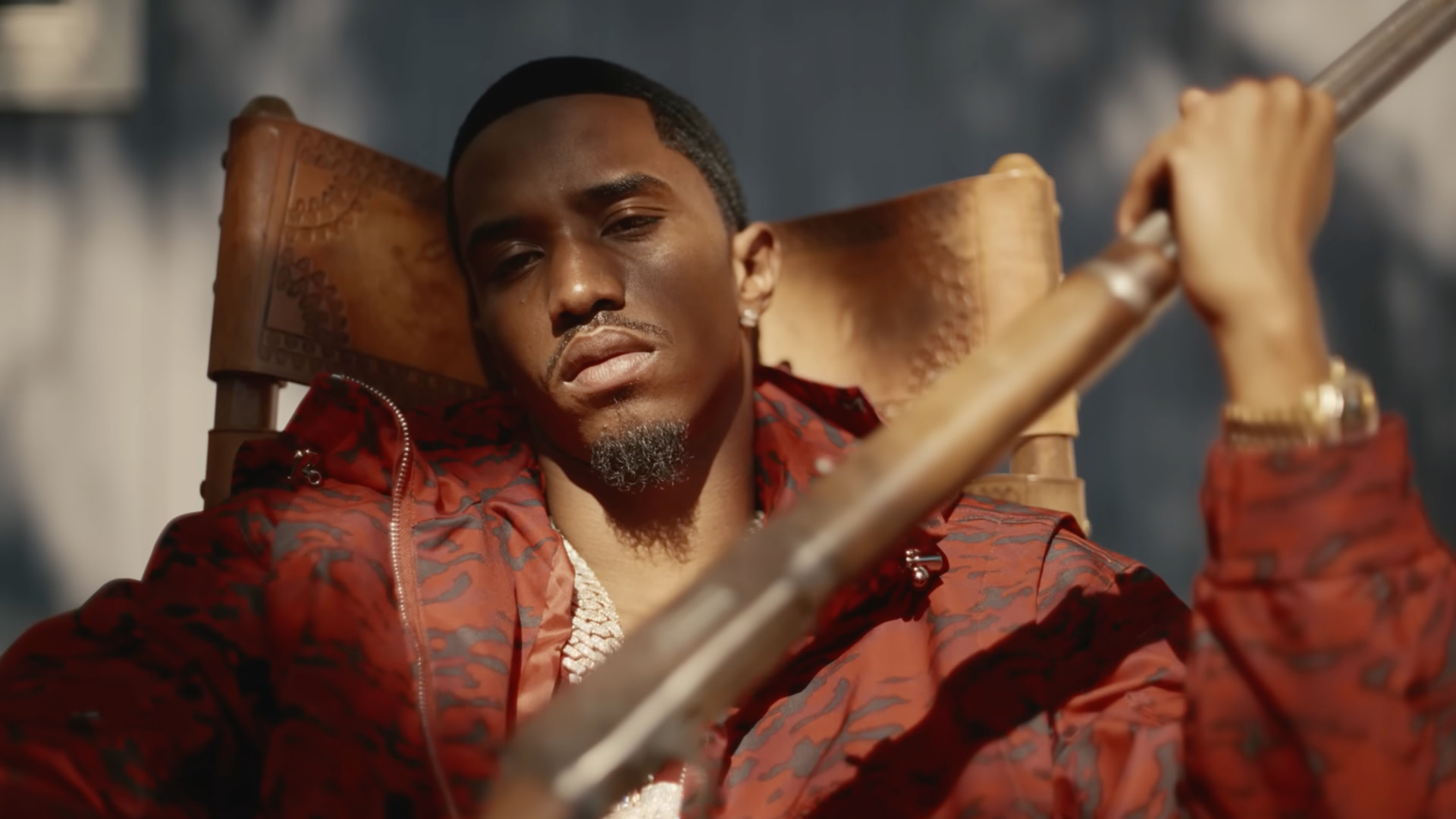 King Combs in a red patterned jacket sits in a chair, holding a long object, with a serious expression.