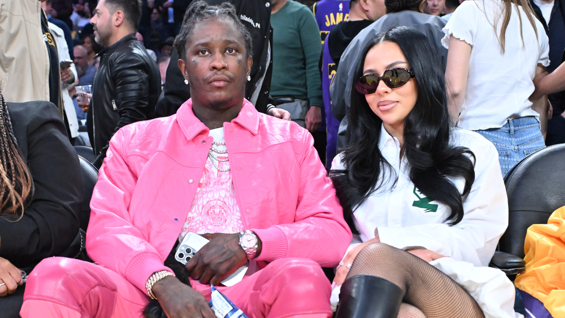 Young Thug in a pink outfit sits courtside next to Mariah the Scientist in sunglasses and a white shirt at a basketball game.