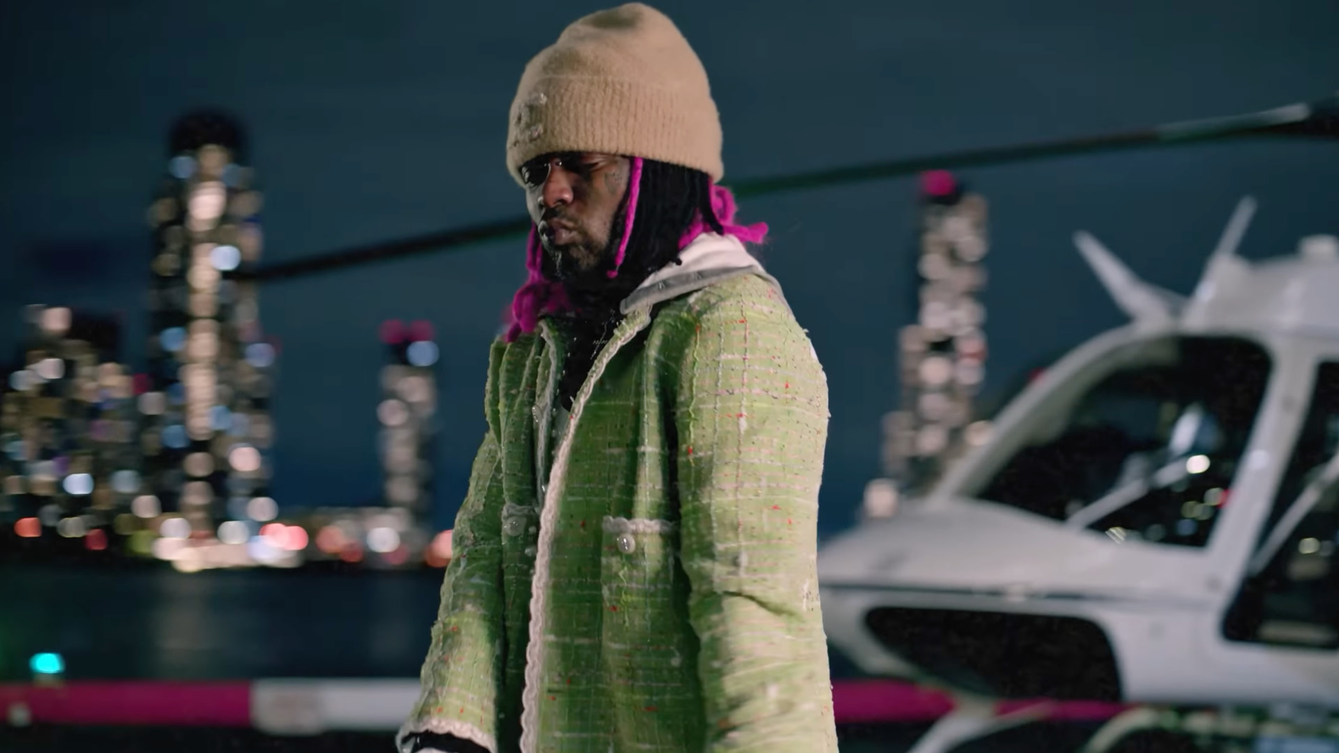 Lil Uzi Vert with pink hair and a green jacket stands near a helicopter, with a city skyline in the background.