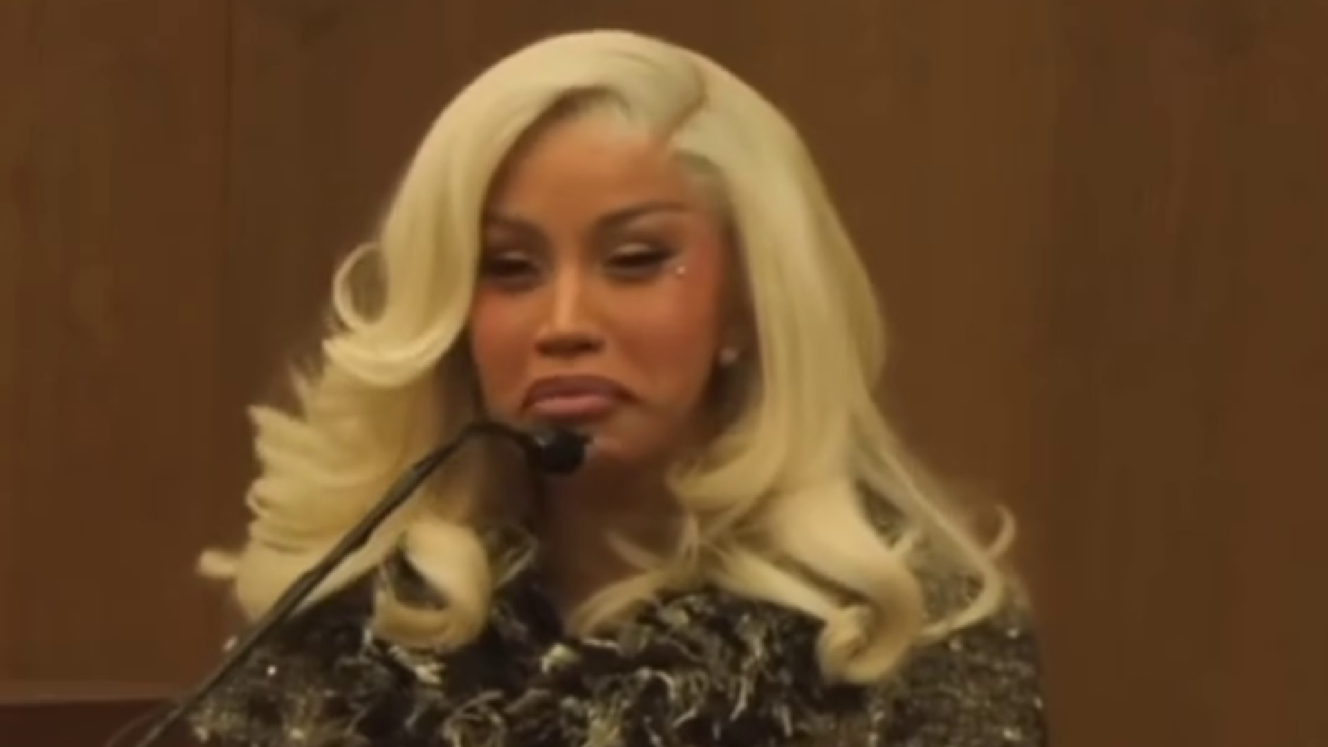 Cardi B with blonde hair, speaking into a microphone, wearing a dark outfit.