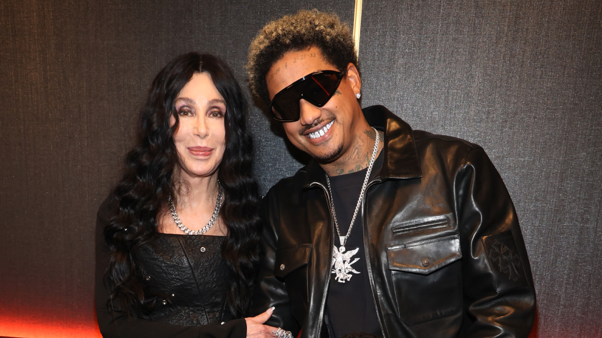 Cher and Alexander Edwards pose together. Cher wears a black outfit and jewelry, while Alexander sports sunglasses and a leather jacket.