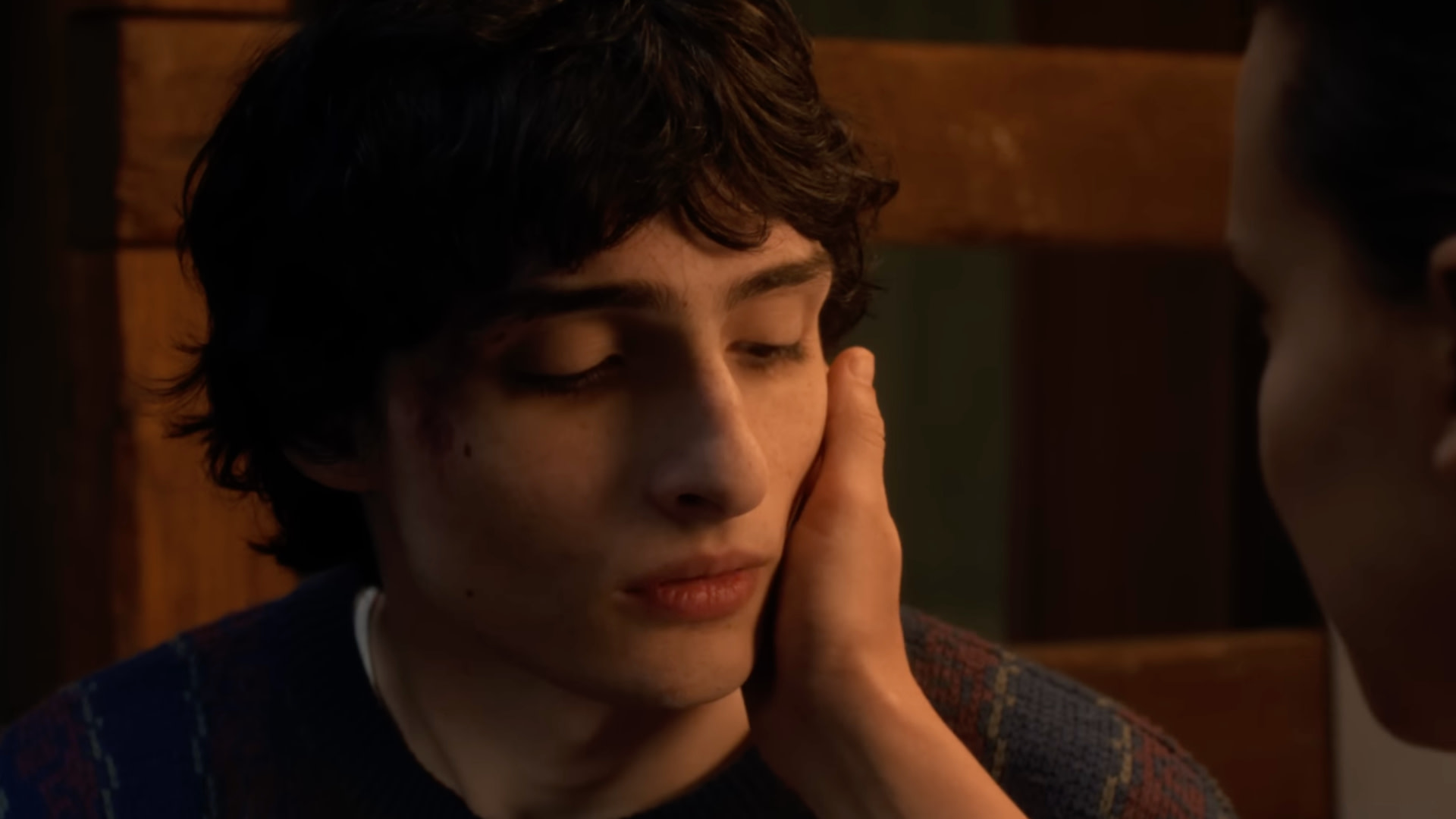 A young person with dark hair, eyes closed, being gently touched on the face by another person. Warm lighting and wooden background.