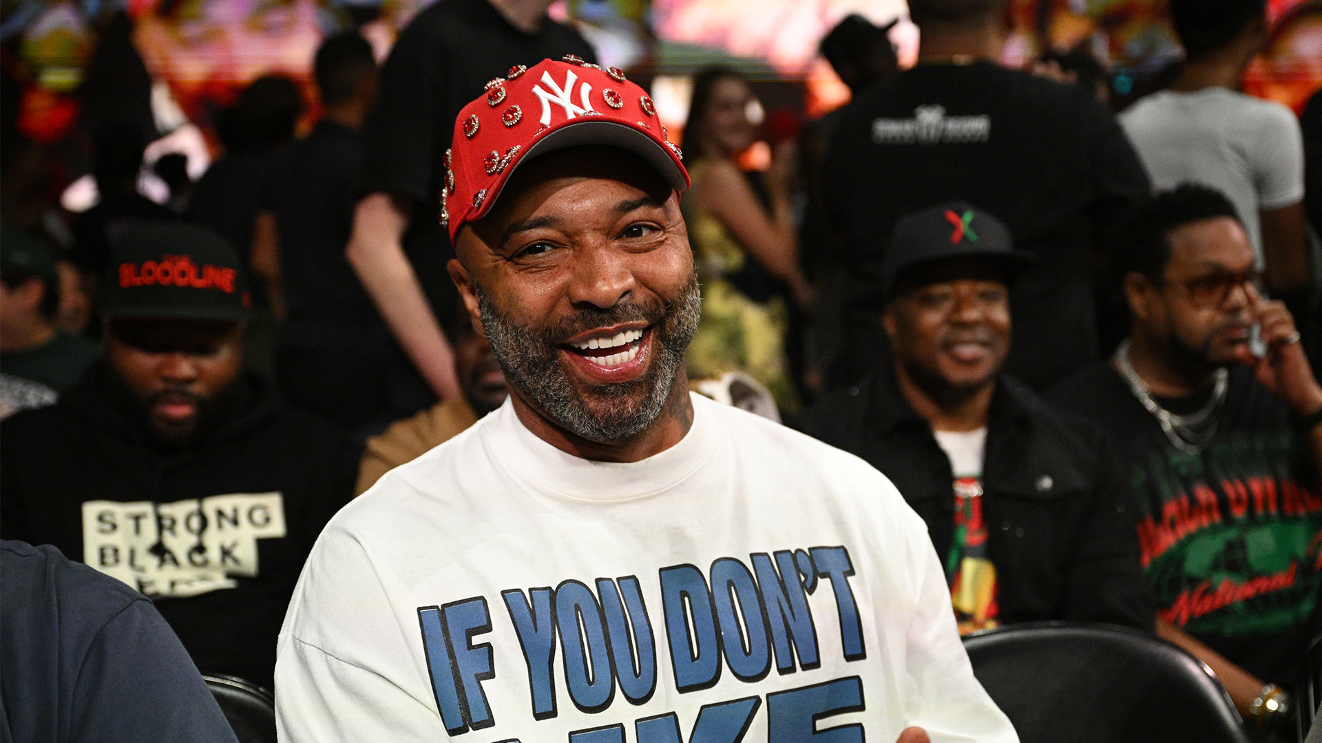 Joe Budden, in a red cap and white shirt with blue text, smiles at an event. People are seated around him in casual attire.