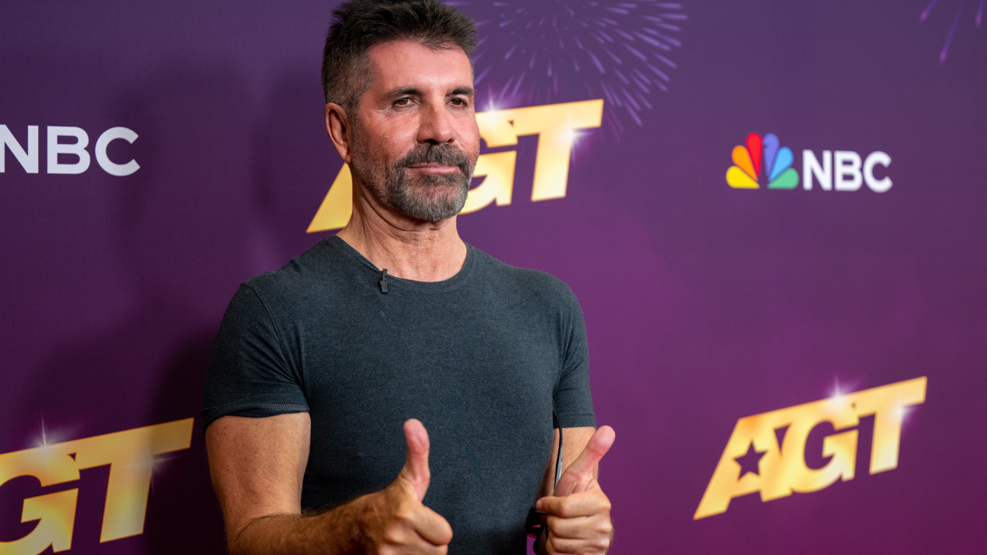 Simon Cowell giving thumbs up on the red carpet at an "AGT" event, wearing a dark T-shirt. NBC logo in the background.