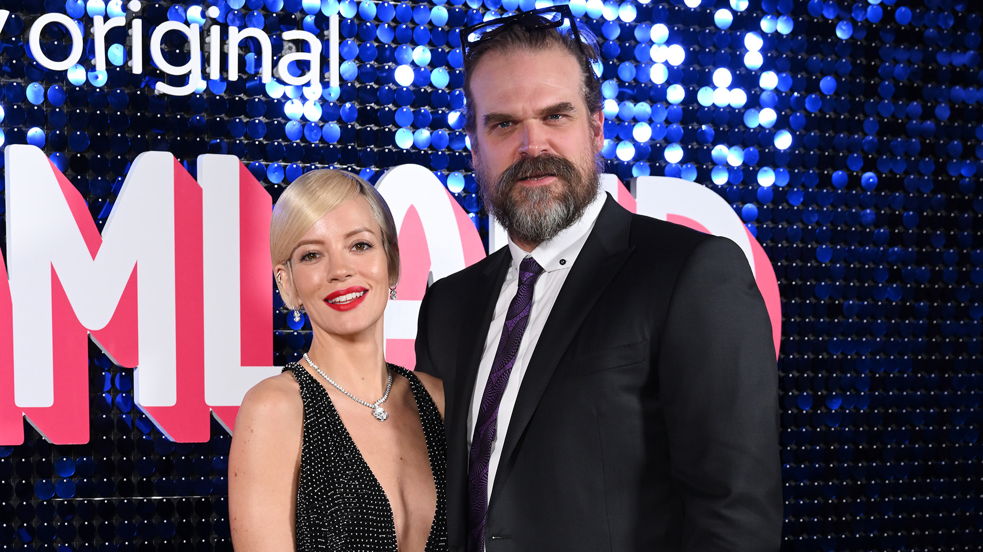 Lily Allen and David Harbour at a red carpet event, with sparkly blue background and "Pamland" signage.
