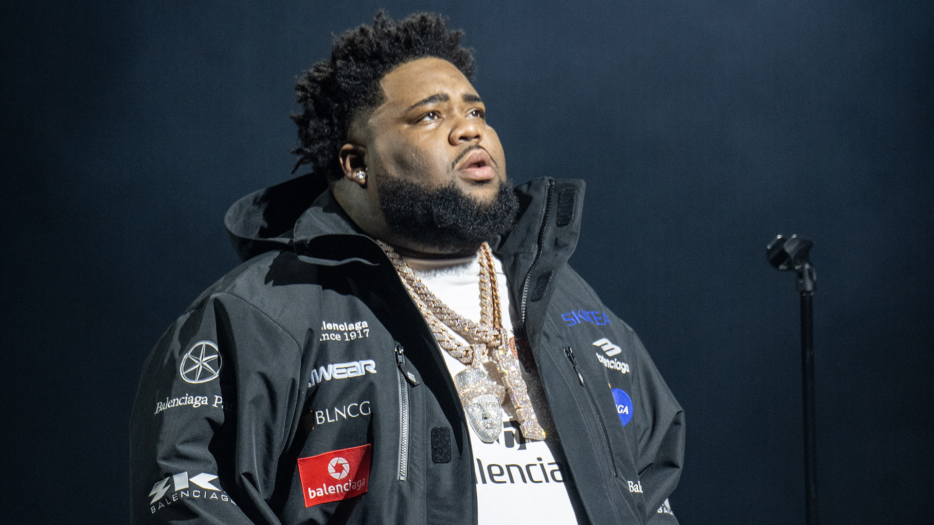 Rod Wave wearing a black jacket with logos, layered necklaces, and a white shirt, stands on stage with a microphone in the background.
